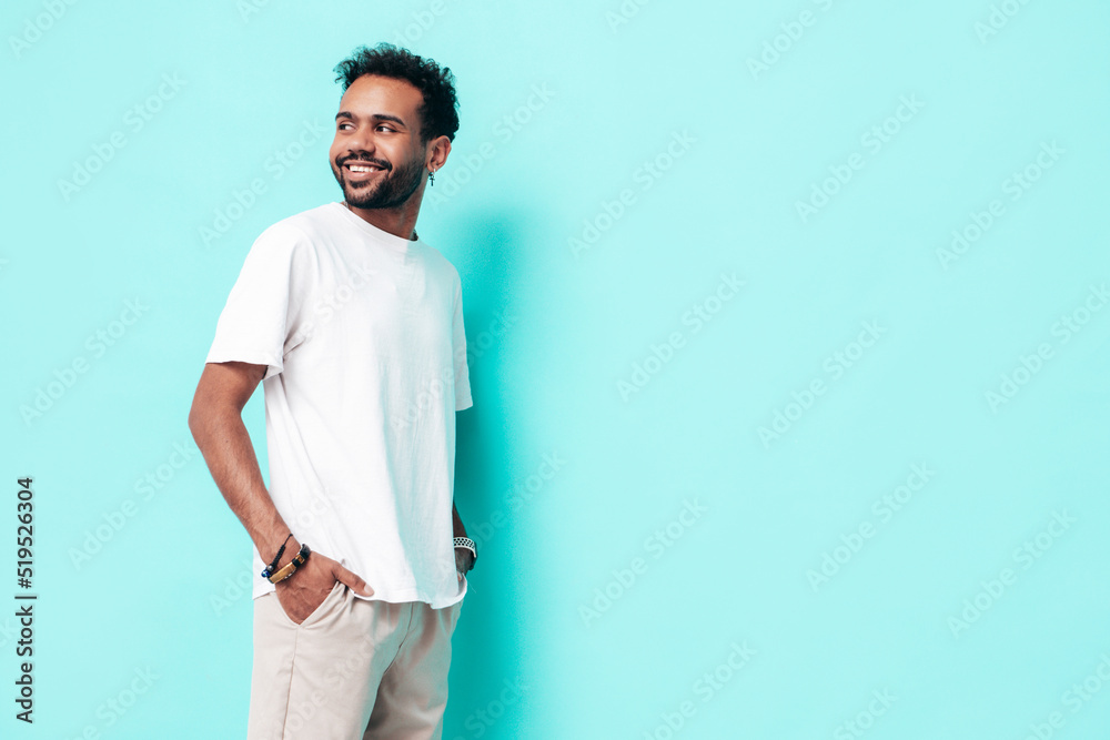 Handsome smiling hipster model. Sexy unshaven man dressed in white summer t-shirt and jeans clothes. Fashion male posing near blue wall in studio. Isolated Handsome smiling hipster model. Sexy unshaven man dressed in white summer t-shirt and jeans clothes. Fashion male posing near blue wall in studio. Isolated