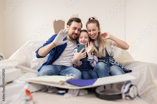 Family dressed in denim style is sitting on the sofa in their new apartment under renovation. All three smilingly wave at the phone camera to their friends with whom they are in online communication.