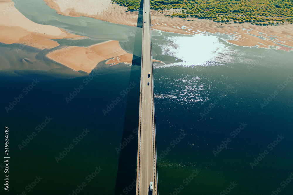 Aerial drone shot of cars driving on bridge over river and sandy riverside at sunset. Amazing aesthetic 4k top view shot with beautiful coast with dune sands. Drone aerial bridge with car traffic