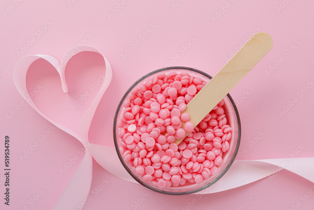 Beautiful granules of pink wax for depilation in a glass bowl, a wooden spatula and a ribbon in the shape of a heart on a pink background. I love waxing. Epilation, depilation, unwanted hair removal. - obrazy, fototapety, plakaty 