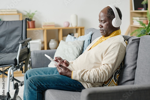 African man with disability sitting on sofa in wireless headphones and using tablet pc for listening music in the room