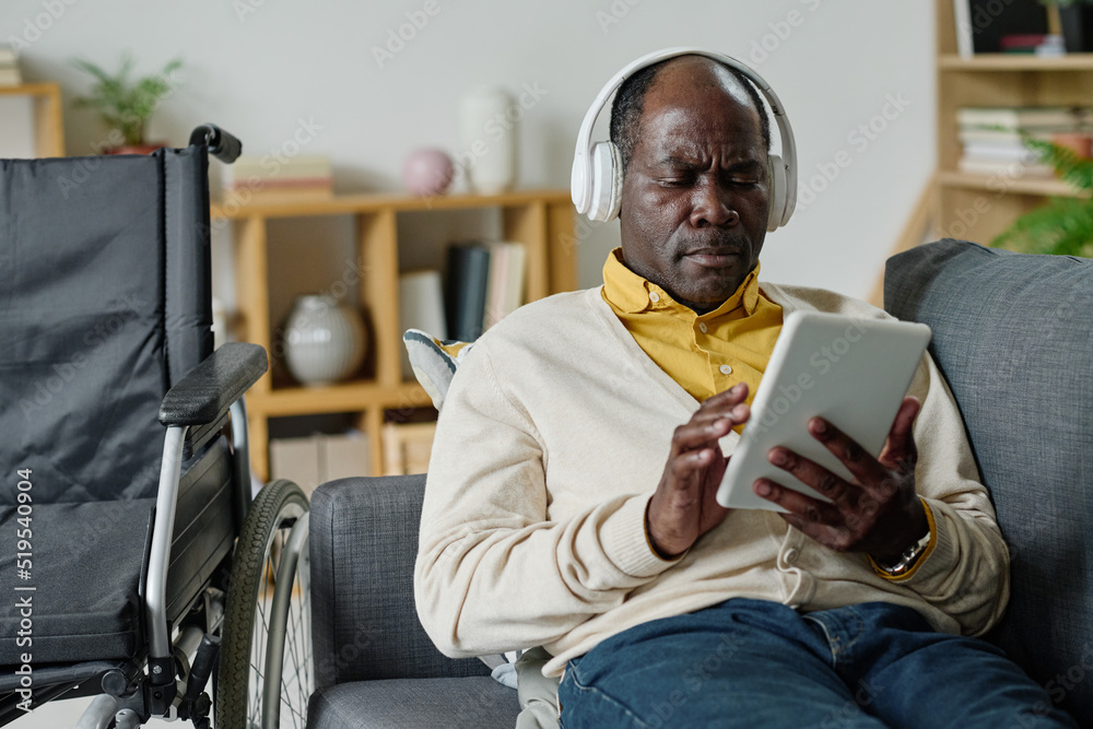 African mature man with disability using digital tablet for musical therapy for relaxation on sofa in living room African mature man with disability using digital tablet for musical therapy for relaxation on sofa in living room