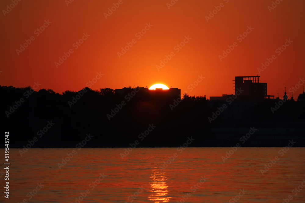 A bright setting sun sets behind the black silhouettes of buildings above the river against an orange red sky. Summer sunset background. Beautiful landscape, close up