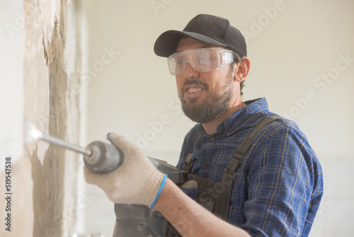 Mature man wearing baseball cap with safety glasses and gloves works on renovation demolition of house uses hammer to tap out tiles in old kitchen bathroom.