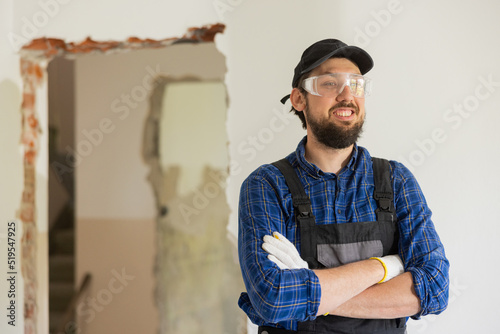 Smiling man in baseball cap wearing protective suit and plaid shirt has goggles on to protect eyes hands crossed over chest stands in middle of renovated house.