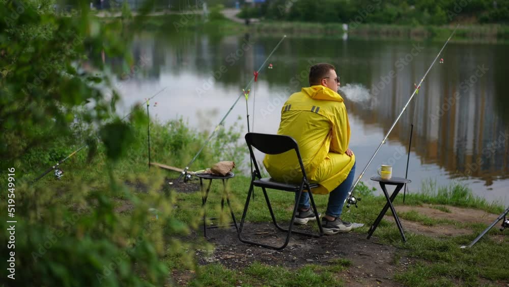 Wide shot young fisherman smoking sitting on river bank waiting for catch. Back view Caucasian man in yellow raincoat and sunglasses enjoying weekend leisure outdoors in park angling. Slow motion
