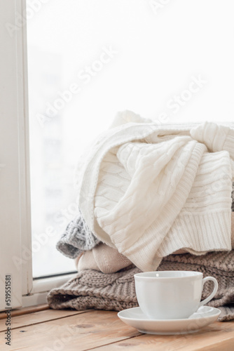 A white cup stands on the windowsill, a pile of warm clothes in the background