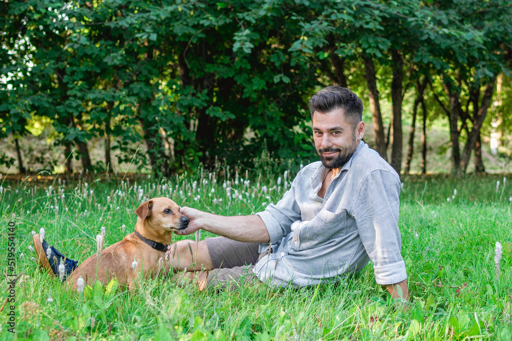 Handsome smiling man sitting on grass with his dog in park. Concept of human and pet relationship. Handsome smiling man sitting on grass with his dog in park. Concept of human and pet relationship.