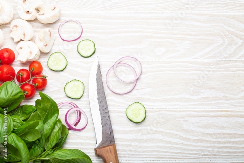 Food cooking background: fresh ingredients with kitchen knife, basil, cherry tomatoes, mushrooms, cut cucumber and onion on white wooden background top view. Meal preparing template, space for text