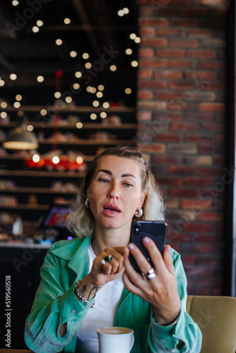 Beautiful happy stylish woman in green shirt sitting in loft cafe  drinking cappuccino  making video call with her cell phone.
