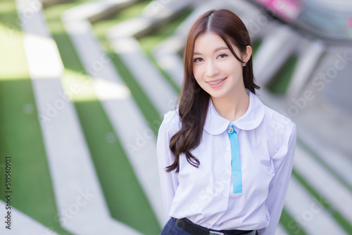 Beautiful high school Asian student girl in the school uniform stands and smiles happily with braces on her teeth while she confidently while she looks at the camera with the building as a background.