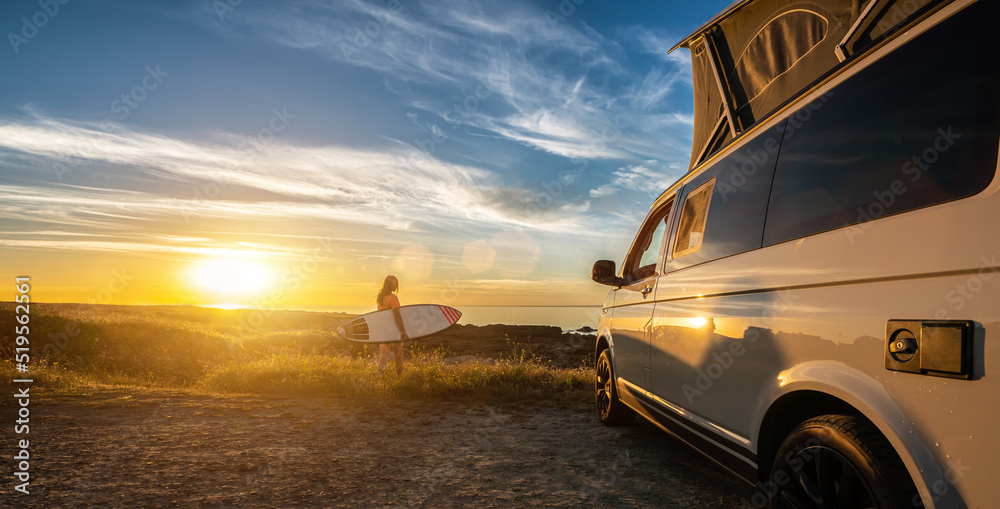 Surfer girl sitting near her mini van and looking on the ocean at summer sunset with a surfboard on her side