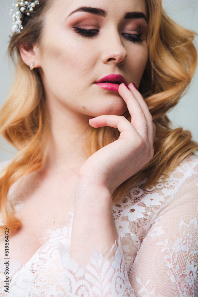 Young attractive bride with blond hair with wedding makeup and hairstyle in a white lace peignoir in a bright interior Young attractive bride with blond hair with wedding makeup and hairstyle in a white lace peignoir in a bright interior
