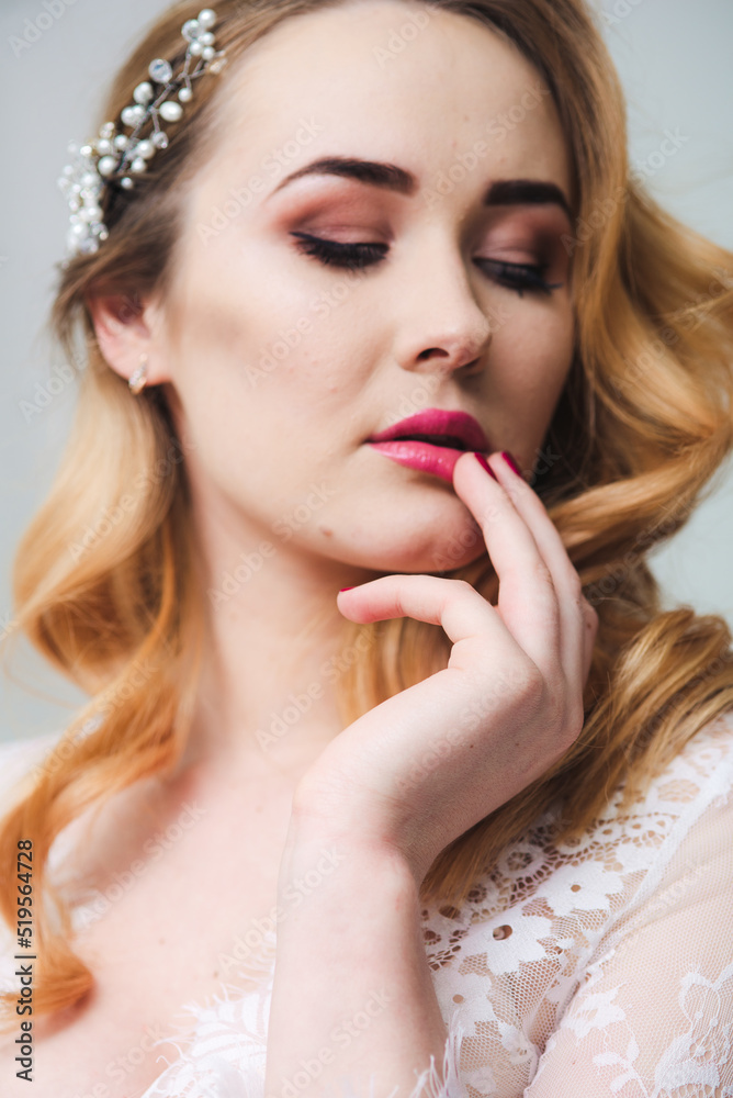 Young attractive bride with blond hair with wedding makeup and hairstyle in a white lace peignoir in a bright interior