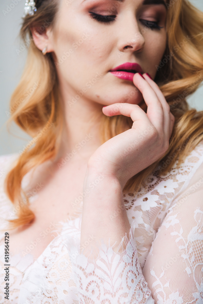 Young attractive bride with blond hair with wedding makeup and hairstyle in a white lace peignoir in a bright interior Young attractive bride with blond hair with wedding makeup and hairstyle in a white lace peignoir in a bright interior
