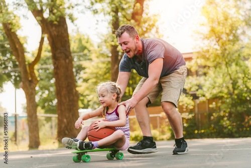 Pretty little girl learning to skateboard outdoors on beautiful summer day with father