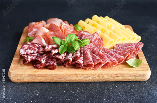 Cheese, prosciutto, salami on a wooden square board on a black stone background. Delicacy.