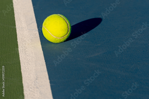 Yellow tennis ball at blue tennis court with white baseline and green out of bounds 