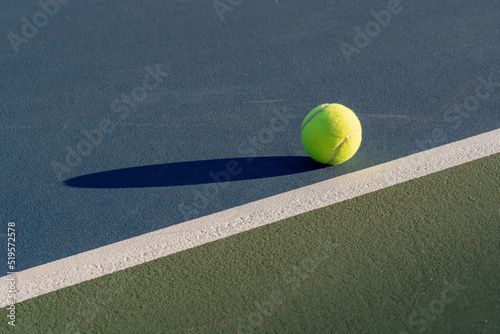 Yellow tennis ball at blue tennis court with white baseline and green out of bounds 