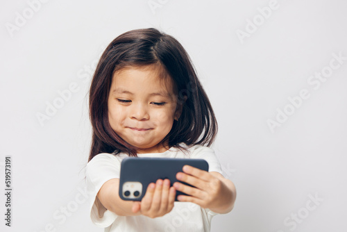 a cute little preschool girl stands on a white background in a white T-shirt and takes pictures of herself on her phone, smiling happily at the camera. The theme of children's happiness