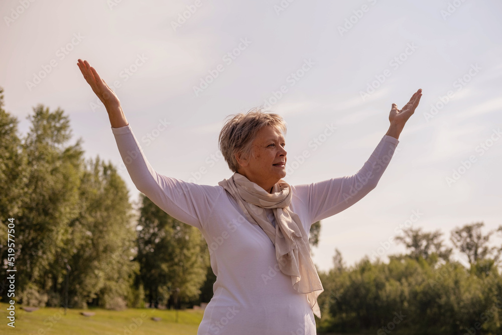 An elderly Caucasian woman stands with her hands raised against the background of green trees and the sky in the park. the concept of success, self-acceptance. An elderly Caucasian woman stands with her hands raised against the background of green trees and the sky in the park. the concept of success, self-acceptance.