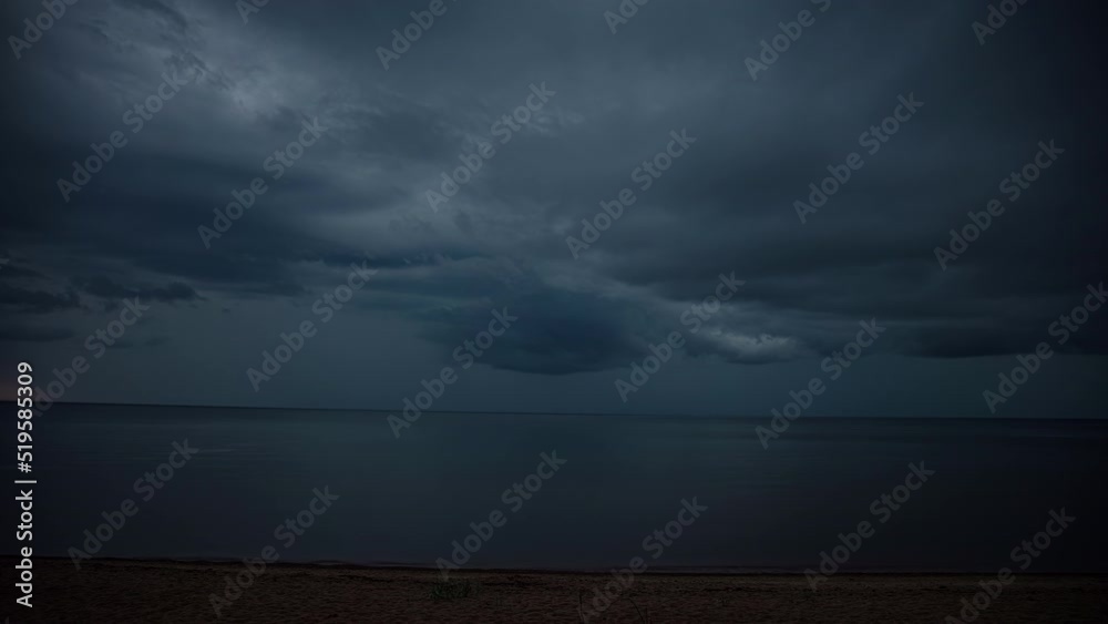 Static view of dark rain clouds passing by over the sea in timelapse along the empty sandy beach. Dark clouds float across the sky during evening time.