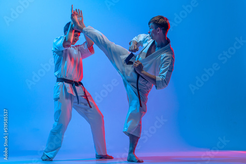 Studio shot of sports training of two karatedo fighters in doboks isolated on blue background in neon. Concept of combat sport, challenges, skills