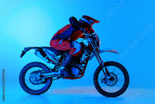 Fototapeta Naklejka Na Ścianę i Meble -  Portrait of young man, biker in full equipment riding motorbike isolated over blue studio background in neon light. Motocross