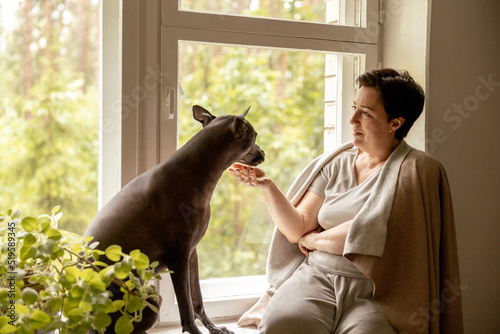 Middle age beautiful woman sitting on windowsill with her dog. 50-year-old woman spending time with her Mexican hairless dog at home. Xoloitzquintle, xolo breed. Dog as best friend, family member. photo