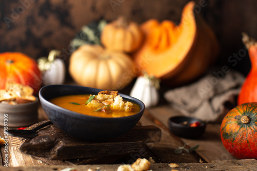 Hot pumpkin soup in ceramic plate with ripe pumpkins,bread croutons garnished with fresh sage leaves on wooden background.