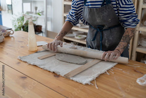 Cropped image of tattooed woman potter master rolling with pin while preparing clay mug, production process of handicrafter pottery. Ceramic making concept