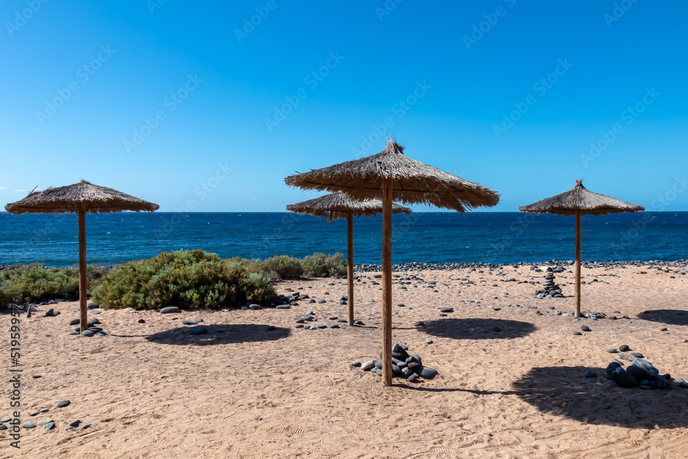 Straw sun umbrellas on sand beach connected to stone pebble beach Playa de San Blas near coastal village Los Abrigos, Tenerife, Canary Islands, Spain, Europe, EU. Coastline of Atlantic Ocean. Vacay