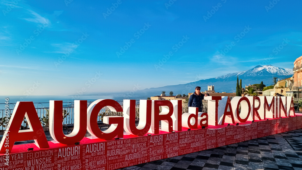 Fototapeta premium Man with christmas tree and signs of Christmas wishes (Auguri) on the main square (Piazza IX Aprile) in Taormina, Province of Messina, Sicily, Italy, Europe, EU. Holidays at the Mediterranean sea
