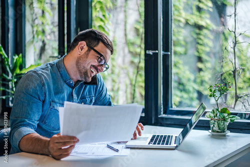 Happy professional with documents talking on cellphone and using laptop at home.