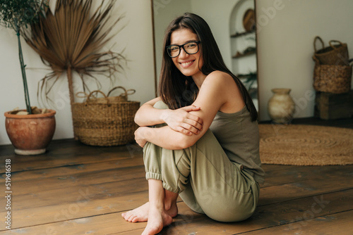 A young twenty year old brunette girl in glasses sits cross-legged on the floor and smiles. photo