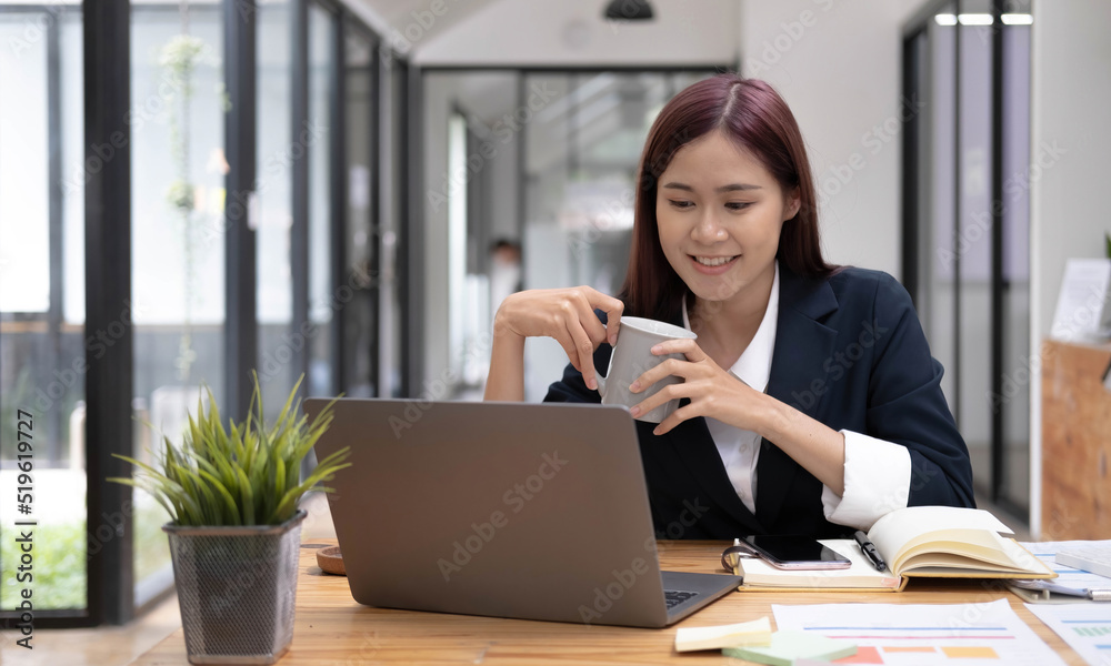 Smiling Asian businesswoman holding a coffee mug and laptop at the office.