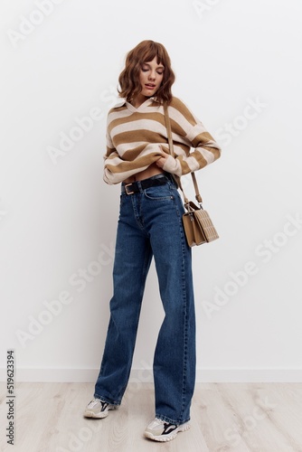 a full-length photo of a beautiful, stylish woman standing on a light background in a striped sweater and blue jeans, posing relaxed with a beige bag worn over her shoulder looking to the side