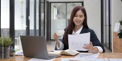 Smiling young Asian businesswoman holding a coffee mug and laptop at the office.