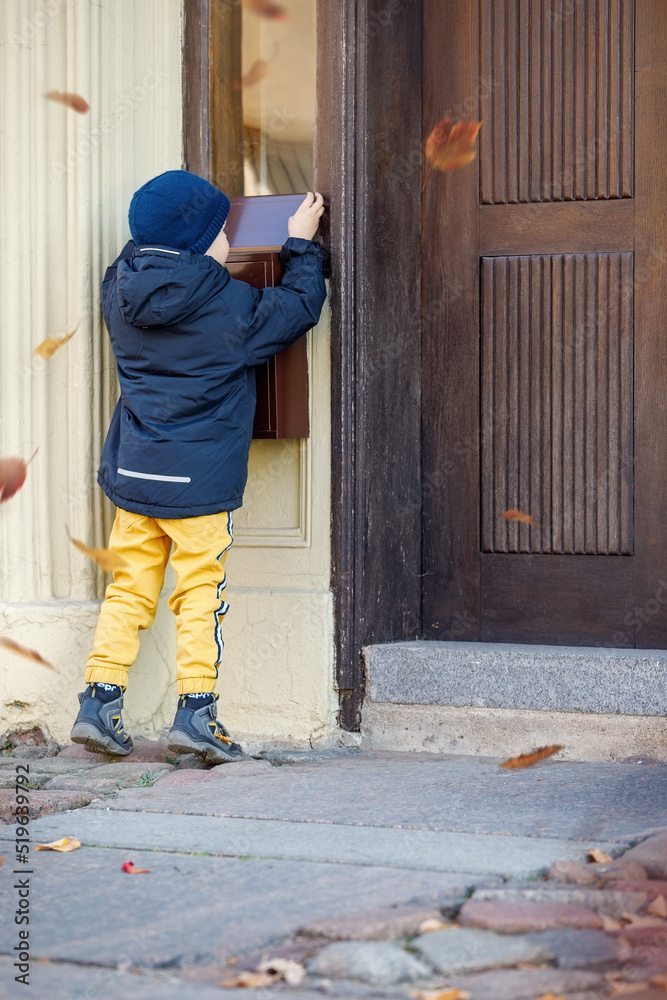 Little boy, checking the mail in outdoors mailbox. The kid is waiting for the letter, checks the correspondence and looks into the metal mailbox. Falling leaves during the autumn season