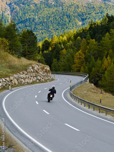 Fotografie Back view of motorcyclist in black jacket and helmet riding sportbike along twisty road between tall green trees and beautiful mountain view