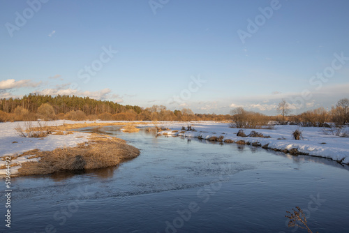 March sunny evening by the river. Blue sky over the horizon. A picturesque landscape  early spring  a river with snow-covered banks  dry grass and bushes. The first thaws  the snow is melting