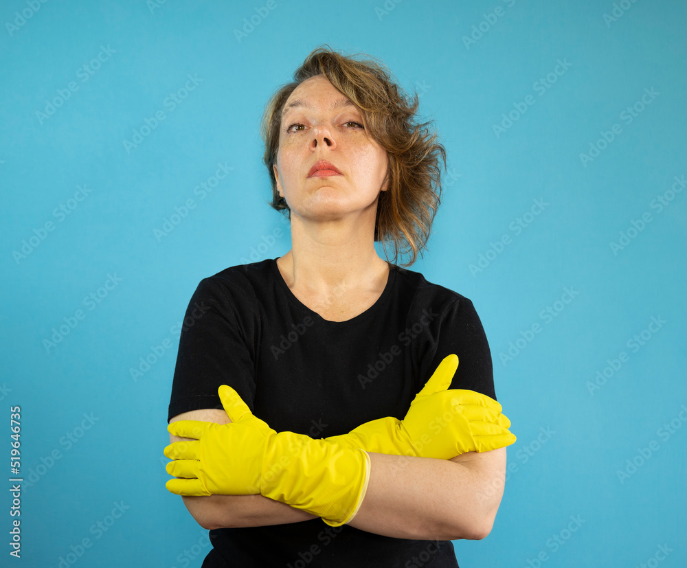 Portrait of a serious cleaning woman in a black t-shirt on an isolated blue background. Woman in yellow rubber gloves for cleaning Portrait of a serious cleaning woman in a black t-shirt on an isolated blue background. Woman in yellow rubber gloves for cleaning