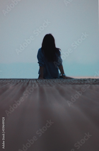 Silhouette of a lonely young girl sitting on a sea pier and looking into the distance beyond the horizon. Loneliness and waiting concept. Evening sunset on the ocean coast. Noisy photo effect