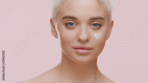 Beautiful young slim light-haired Caucasian woman with white cream on her cheek smiles for the camera looking at it on pale pink background   Face care cream commercial