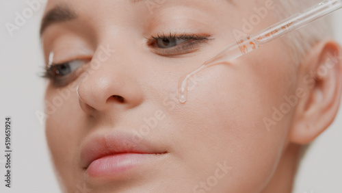 Cute young slim fair-haired Caucasian woman drips transparent serum from dropper on her cheek on white background | Skin care serum commercial