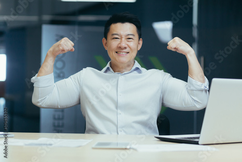 A successful and handsome young Asian business man looks at the camera, smiles, shows with his hands that he is strong. Sitting at the desk in the office, celebrating victory, success