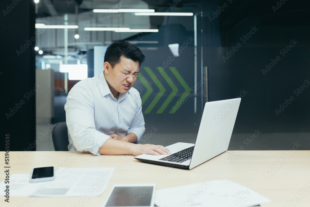 A young Asian businessman holds his stomach at the desk in the office, feels bad, feels pain, needs help. A young Asian businessman holds his stomach at the desk in the office, feels bad, feels pain, needs help.