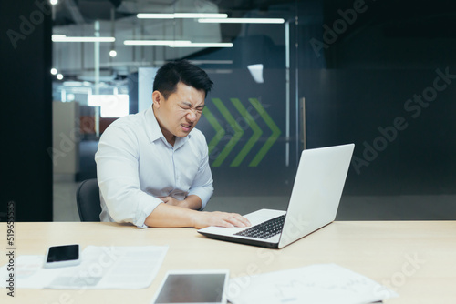 A young Asian businessman holds his stomach at the desk in the office, feels bad, feels pain, needs help.