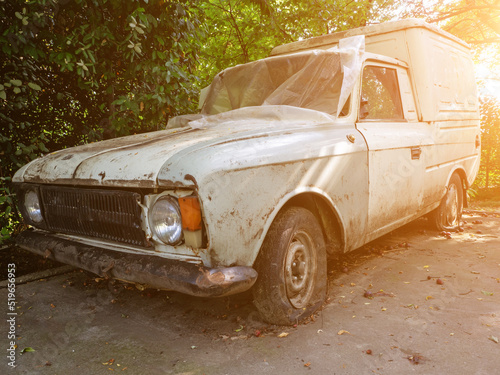 Obsolete dirty car with flat tires on the street. A dirty white cargo-passenger passenger car of an outdated model with flat wheels stands in the yard covered with polyethylene photo