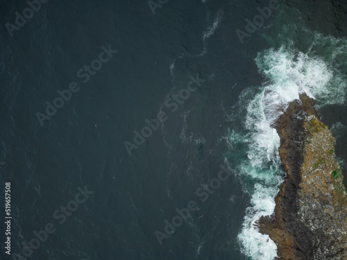 Shooting from a drone. Dark turquoise ocean surface. Foamy waves crash against the rocky shore. Minimalism. There are no people in the photo. Advertising tourist routes, ecology. © Anton